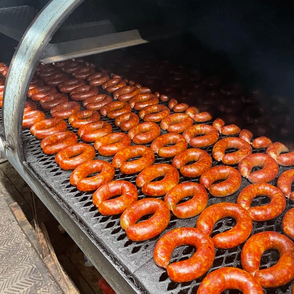 Rows of reddish-brown sausages cooking on a large, dark grill inside a smoker.