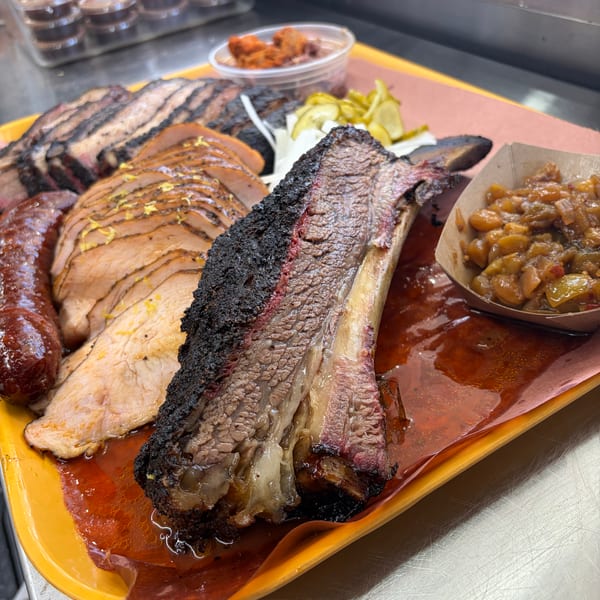 A platter of smoked meats including brisket, sausage, sliced turkey, and sides of pickles and beans.