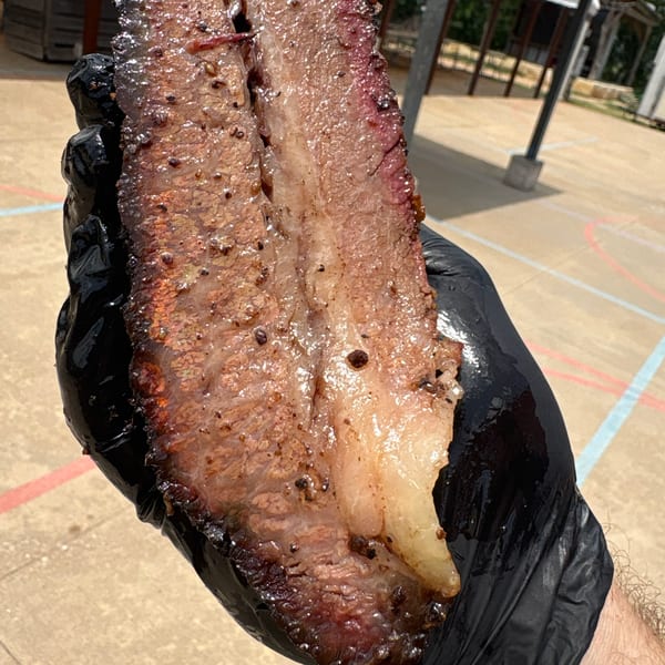 A gloved hand holds a large, glistening slice of smoked brisket with a prominent smoke ring.