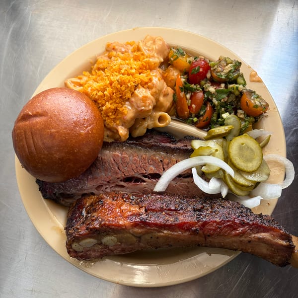 A plate of BBQ brisket and ribs with mac and cheese, tomato salad, pickles, and a bun.